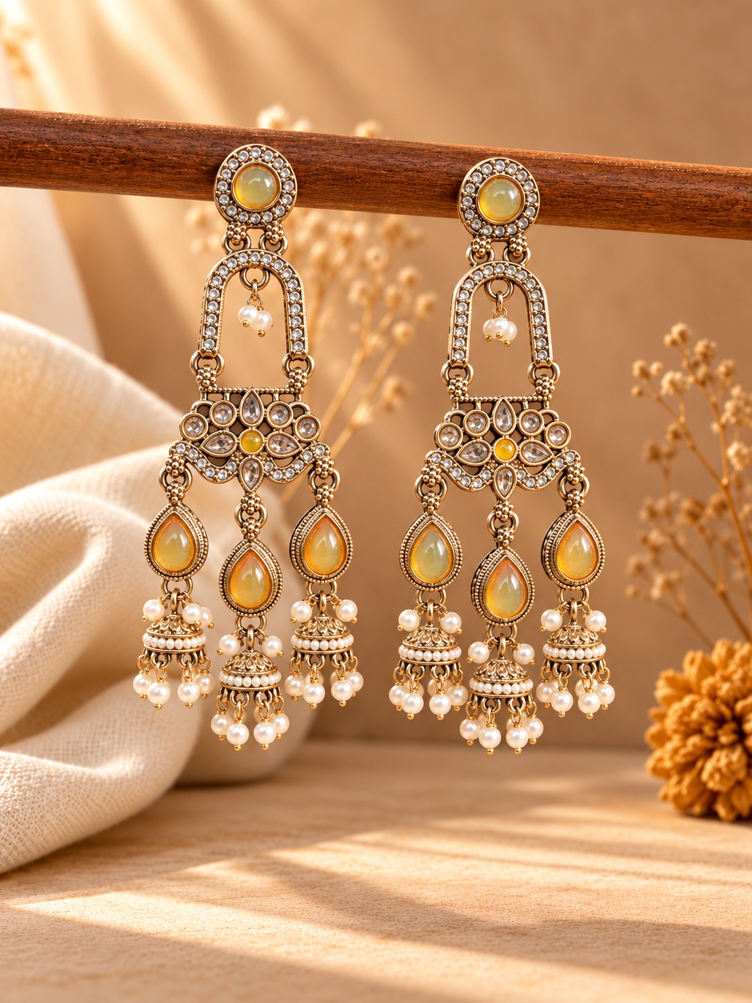 Mrinalika yellow triple jhumka earrings with floral stones and white bead fringes hanging from a wooden rod